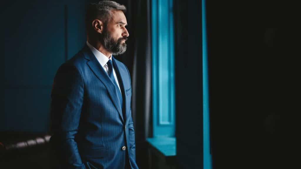 A bearded man in a blue suit looks out a window in a dimly lit room.
