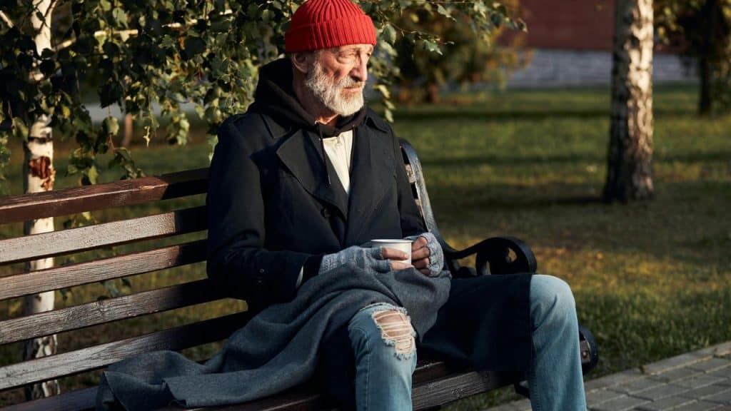 An elderly man in a red beanie sits on a park bench holding a cup.
