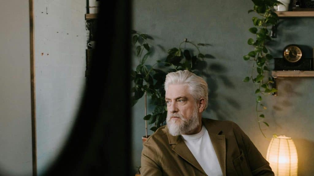 An older man with white hair and a beard sitting indoors near plants and a lamp