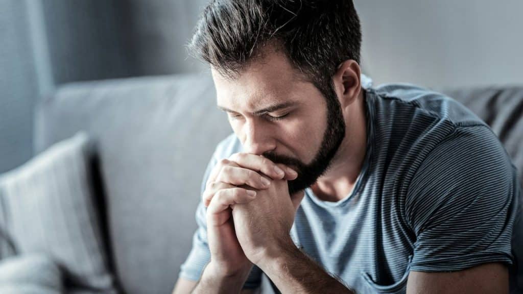 A bearded man in a striped shirt sits on a sofa with his hands clasped.