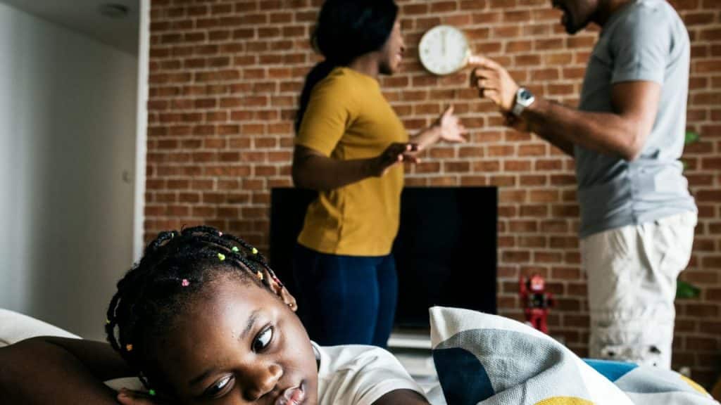 A young girl looks toward the camera while two adults argue in the background indoors.