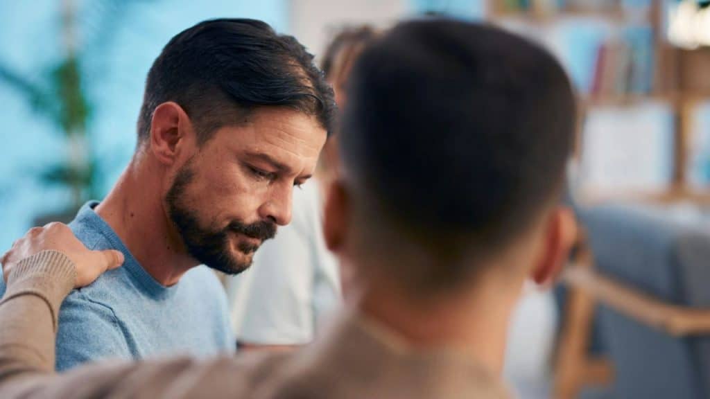 A man with a beard looks down as another person places a hand on his shoulder.