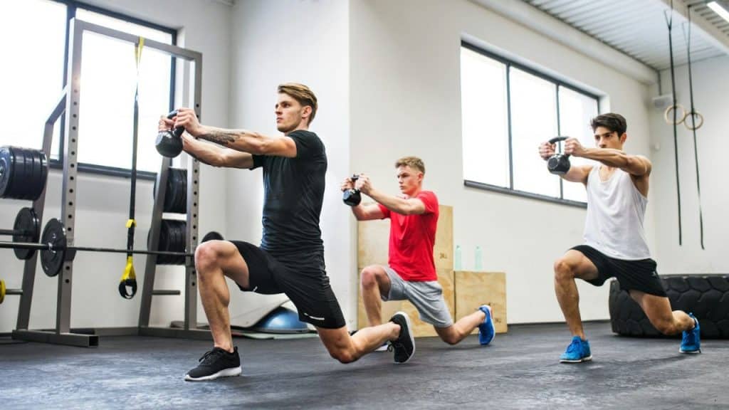 Three men in athletic wear perform lunges while holding kettlebells in a bright gymnasium.