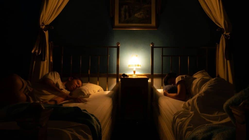 Two people sleep in separate twin beds divided by a nightstand with a lit lamp.