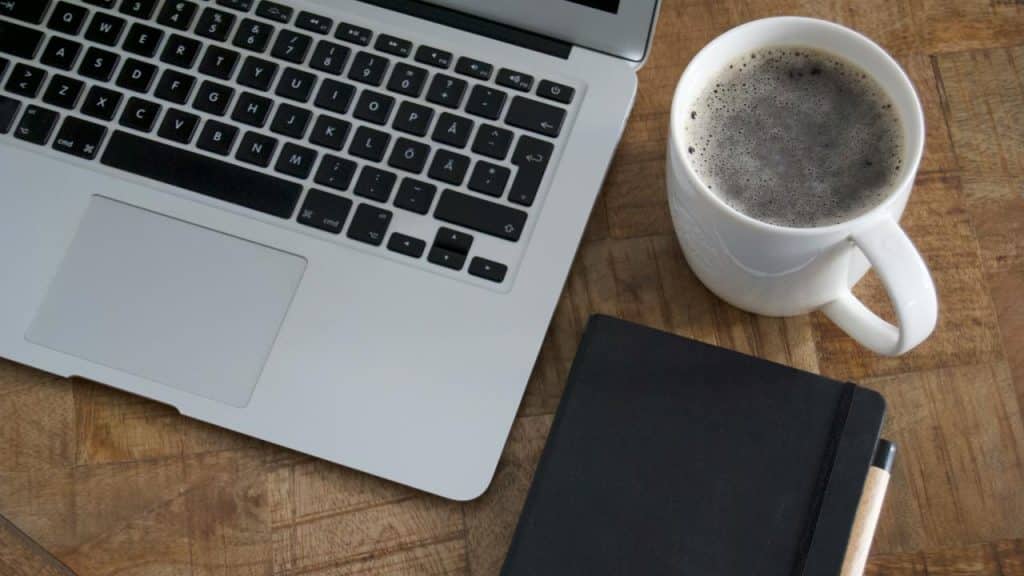 A laptop, coffee mug, and notebook on a wooden desk.