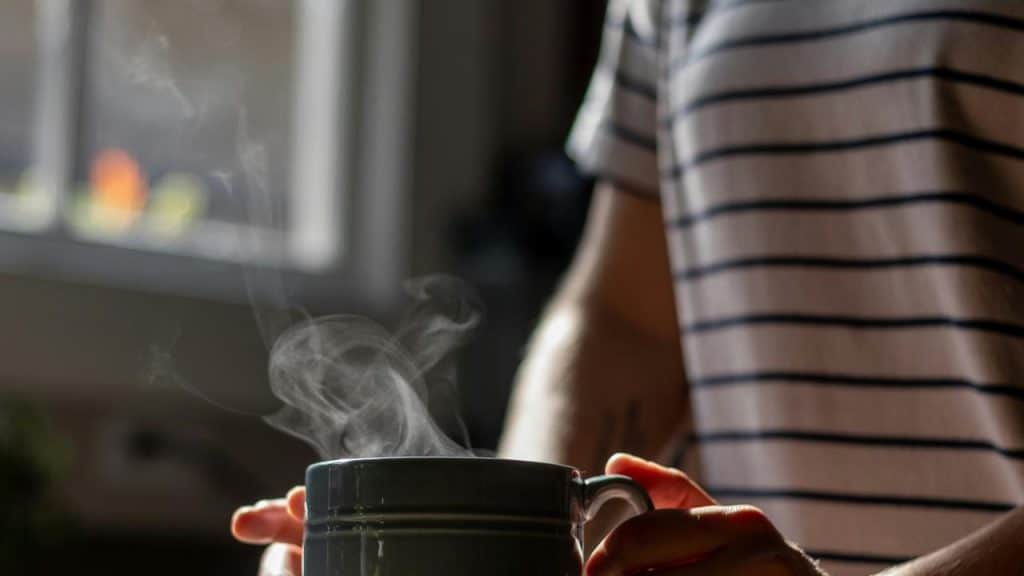 A person holds a steaming mug near a window.
