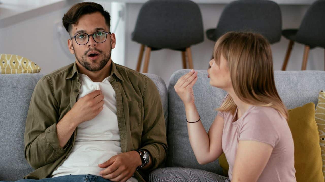 A man with glasses looks surprised while a woman gestures toward him on a sofa.