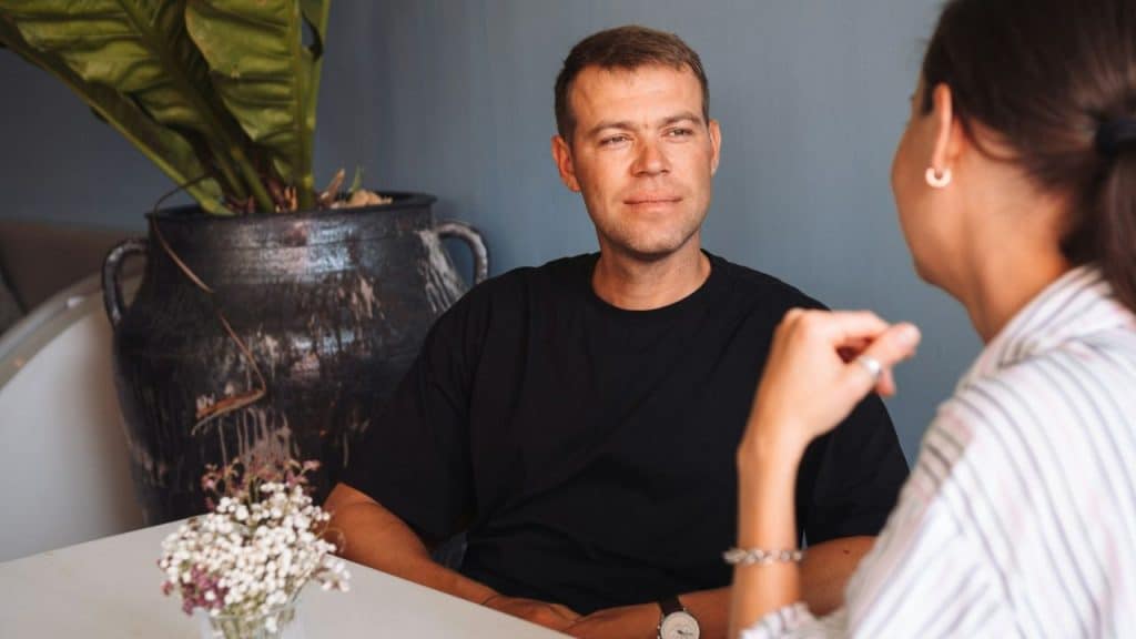 A man in a black shirt looks at a woman across a table, with a plant in a large pot in the background.