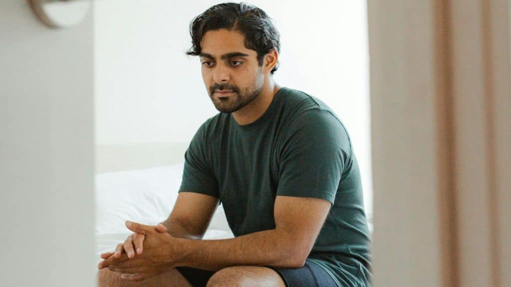A man with a beard and dark hair sitting indoors, looking down with hands clasped.