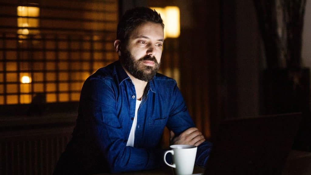 A bearded man in a blue shirt sits at a desk, looking at a laptop.