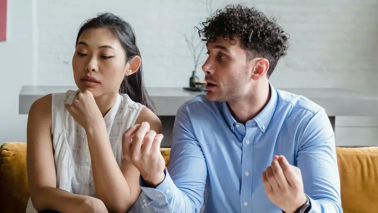 A man with curly hair gestures with both hands while speaking to a woman looking away.