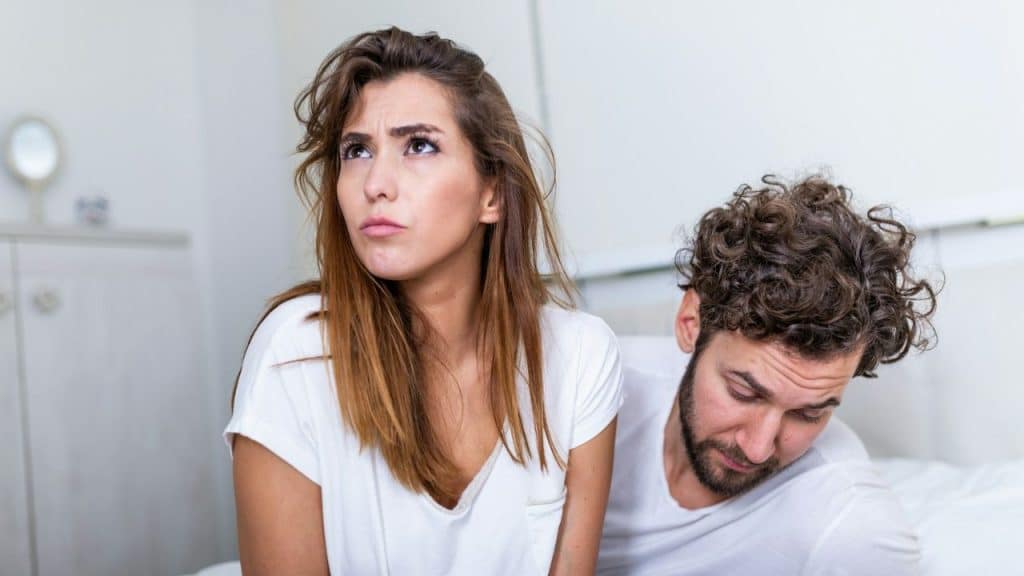 A woman looks upward with a frustrated expression while a man sits beside her looking down.