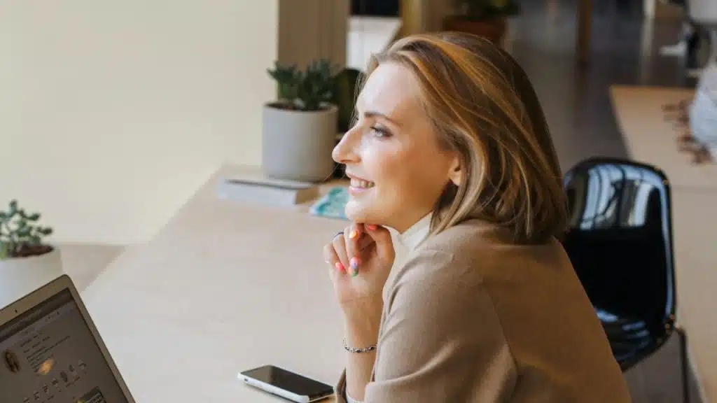 A smiling woman in a brown blazer sits at a desk with a laptop and phone.