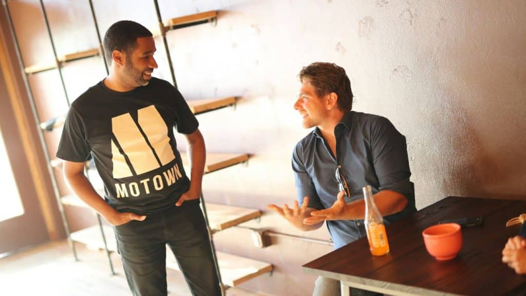 Two men in casual clothing stand and sit in a room, smiling and gesturing while talking.
