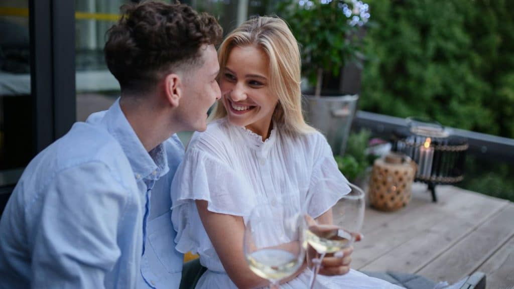 A man and woman sit on a deck, smiling at each other while holding wine glasses.