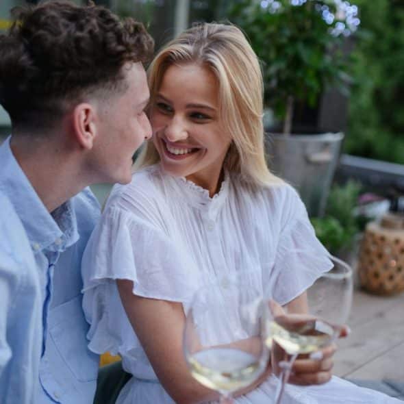 A man and woman sit on a deck, smiling at each other while holding wine glasses.