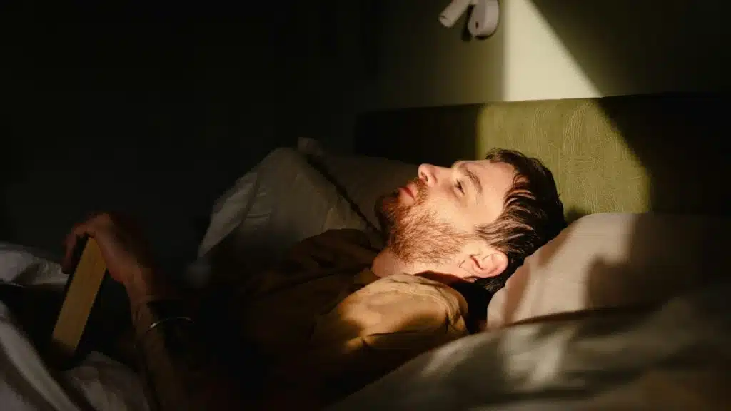 A man with a beard lies in bed, holding a book while illuminated by sunlight.