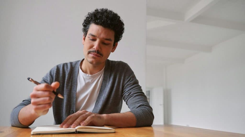 A man with curly hair and a mustache writing in a notebook at a table.