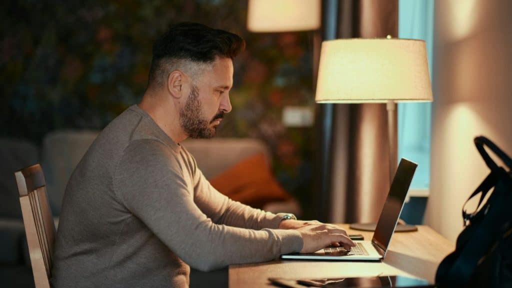 A man with a beard sits at a desk working on a laptop near a lamp.
