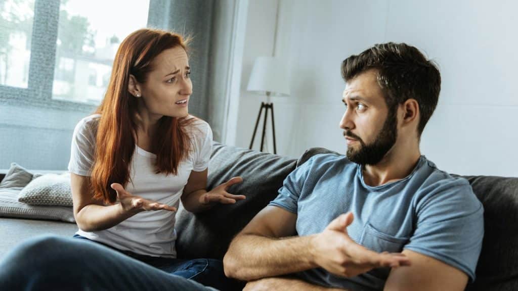 A woman arguing with a bearded man who gestures with his hand as they sit on a couch.