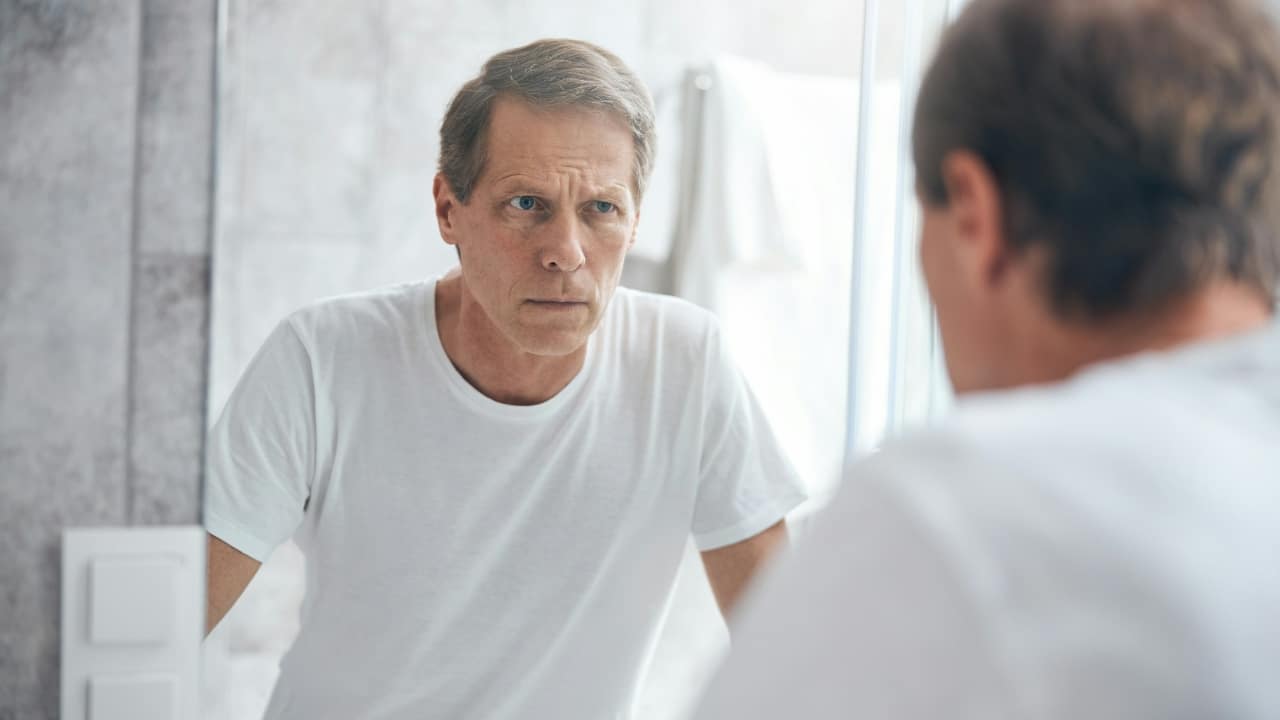 A man in a white t-shirt looks at his reflection in a bathroom mirror.