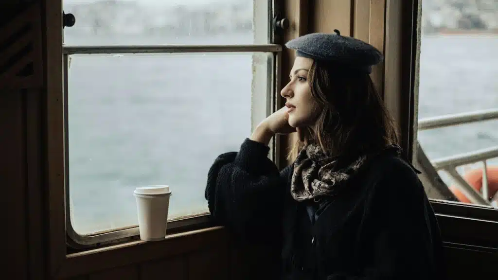 A woman wearing an artist cap and staring out of a train’s window at a body of water.