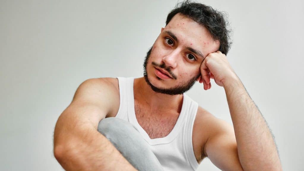A man in a white tank top sits with his head resting on his hand, looking tired.