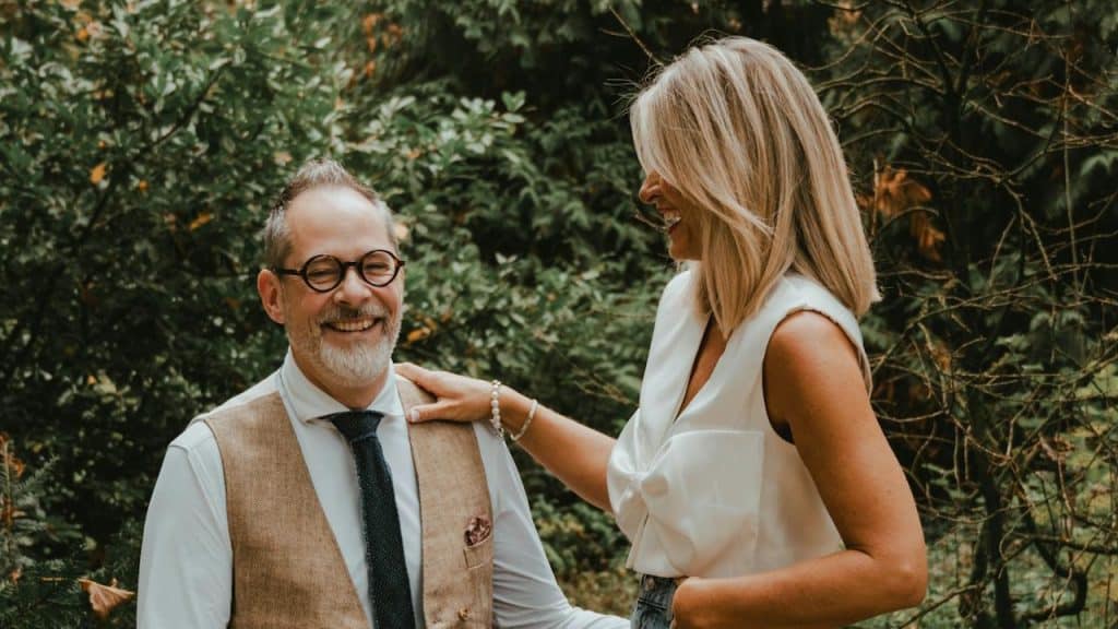A laughing man in a vest and tie sits on a stone wall while a woman in a white top stands beside him, resting her hand on his shoulder in a lush, wooded setting.
