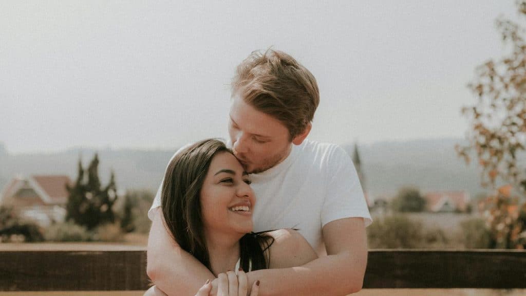A couple embracing outdoors while smiling as one kisses the other’s head.