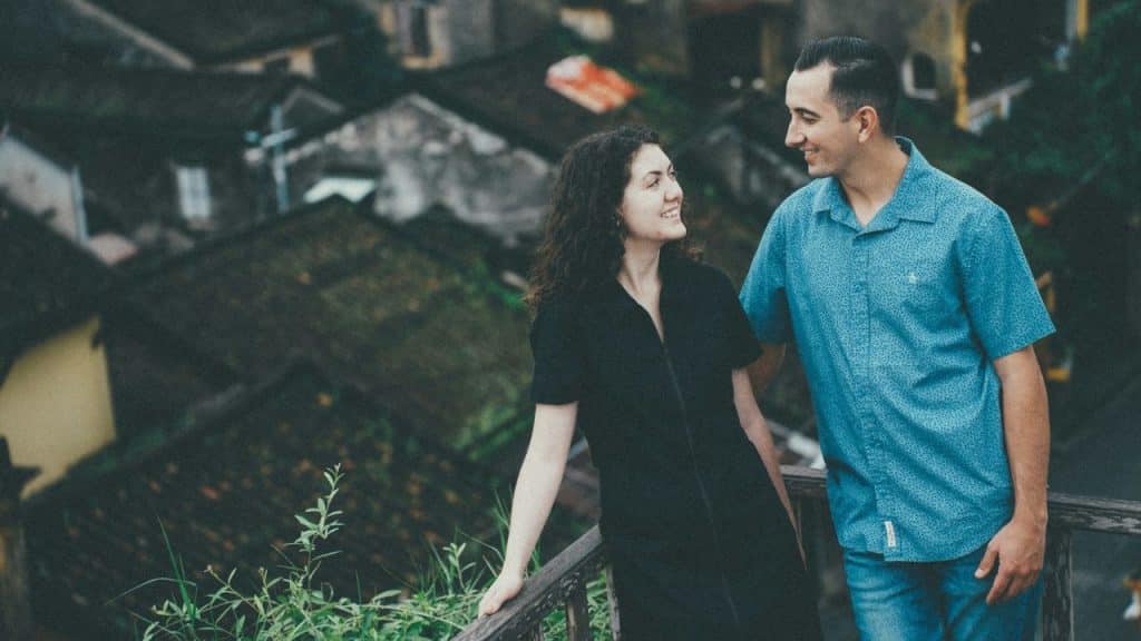 A man and woman smile at each other while standing on a wooden balcony overlooking rooftops.