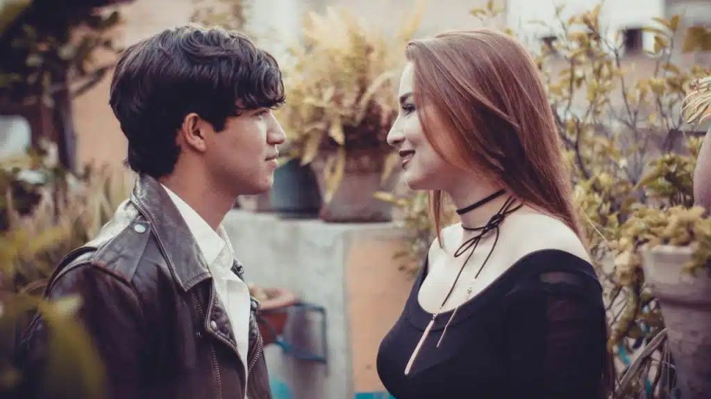 A man and woman face each other outdoors, making eye contact while surrounded by plants.