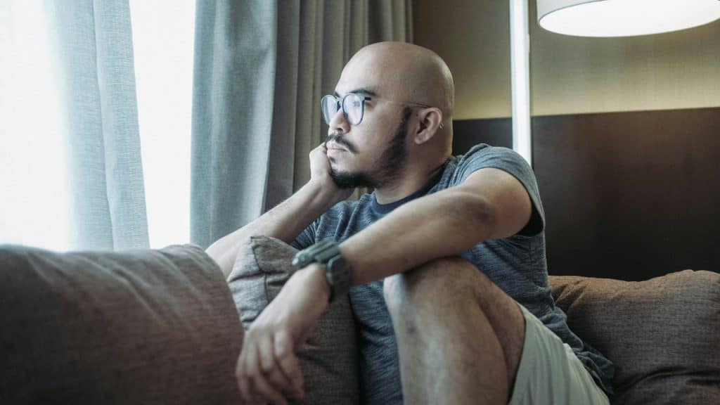 A bald man wearing glasses sits on a couch, resting his chin on his hand.