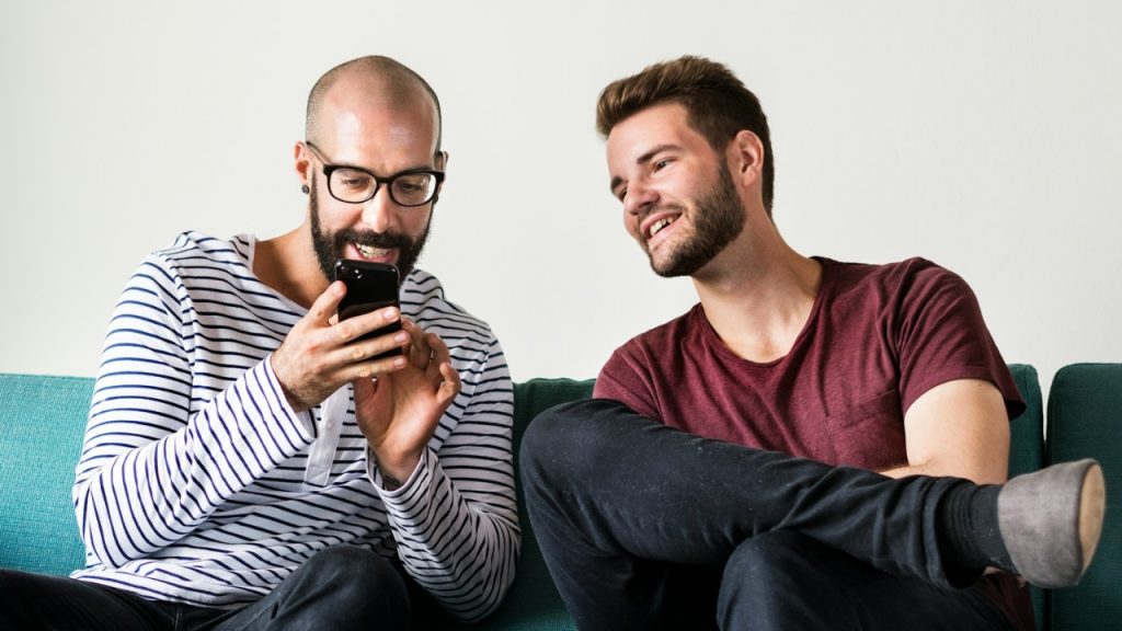 Two men sit on a green sofa looking at a smartphone and smiling.