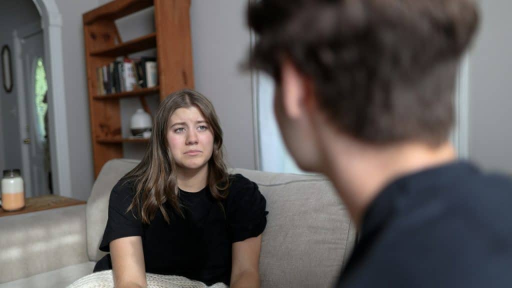 A woman with a sad expression sits on a couch looking at a blurry man.