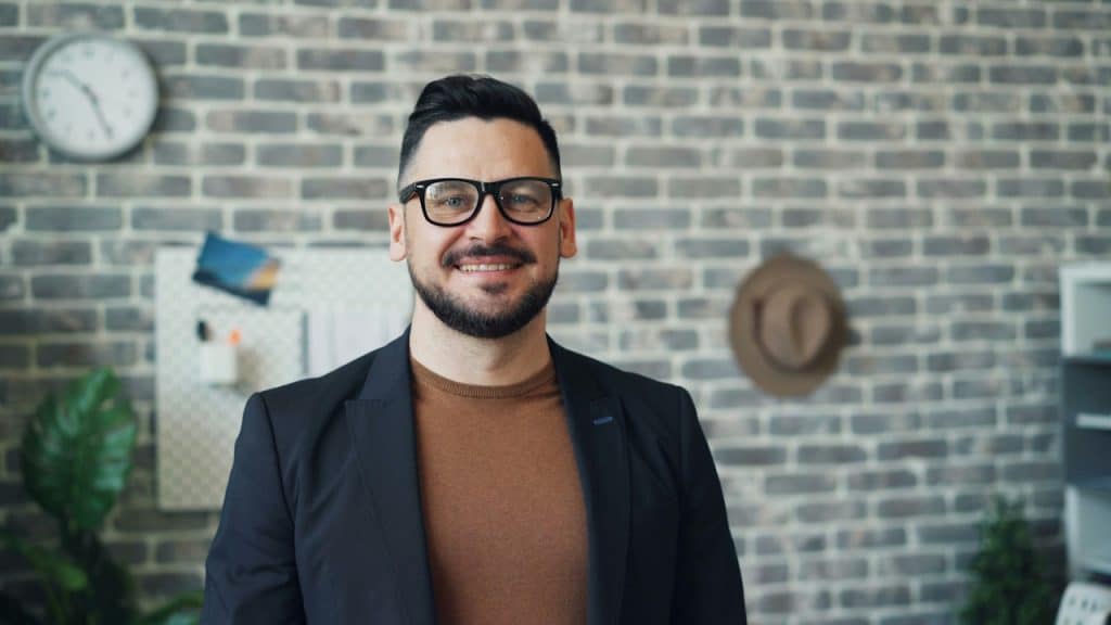 A man wearing glasses standing in front of a brick wall.
