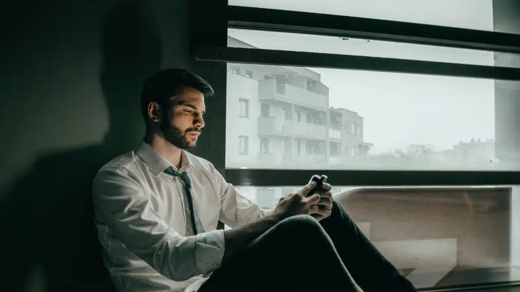 A pensive man sitting by a window and looking at his phone.