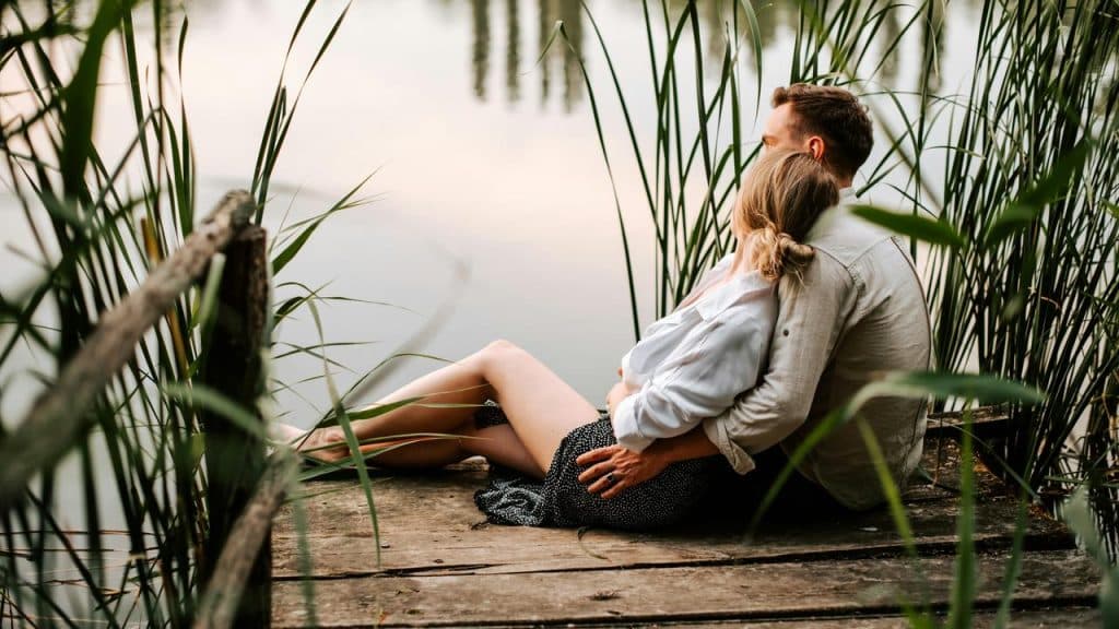A couple sitting on a wooden dock by a lakeside reeds.