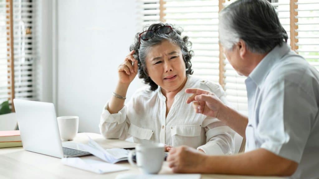 A concerned woman with gray hair listens to a man gesturing, seated at a table with a laptop.