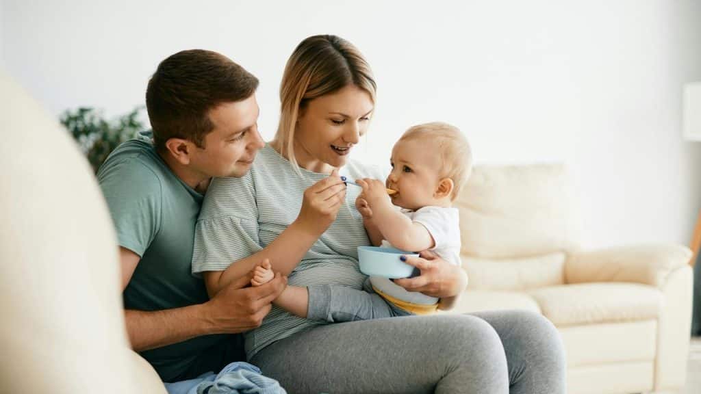 A mother feeds a baby while the father smiles beside them on a couch.