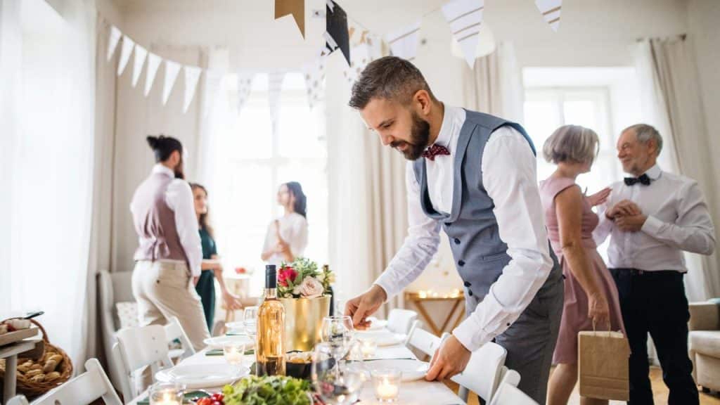 A man in a gray vest setting a decorated table at a wedding reception.