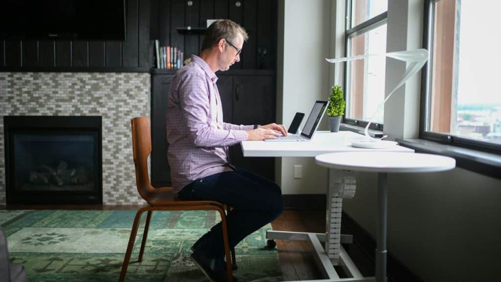 A man in a patterned shirt sits at a white desk working on a laptop.