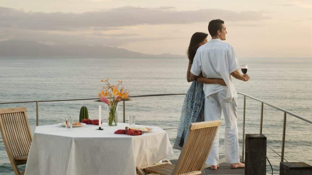A couple stands on a balcony overlooking the ocean next to a set dinner table.