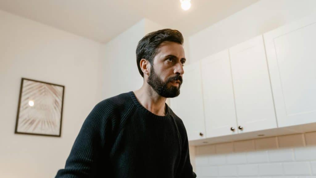 A bearded man standing indoors, looking to the side.