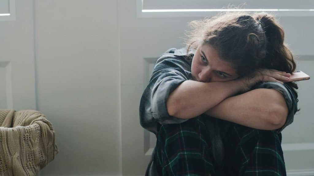 A young woman sitting on the floor hugging her knees and looking distressed.