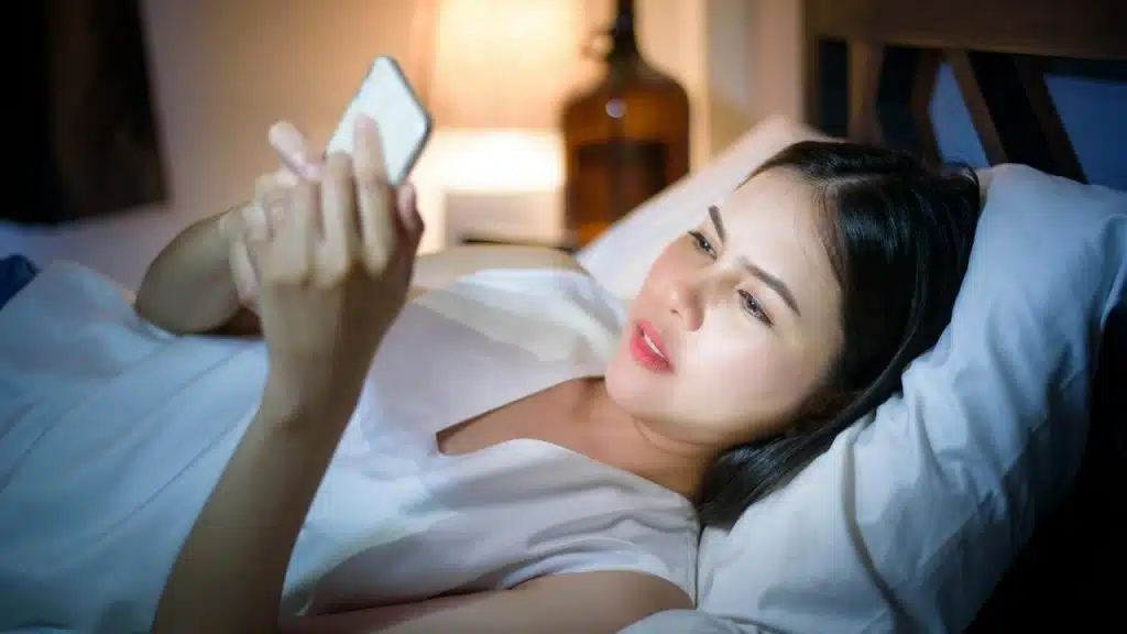 A woman lies in bed at night, looking at her phone.