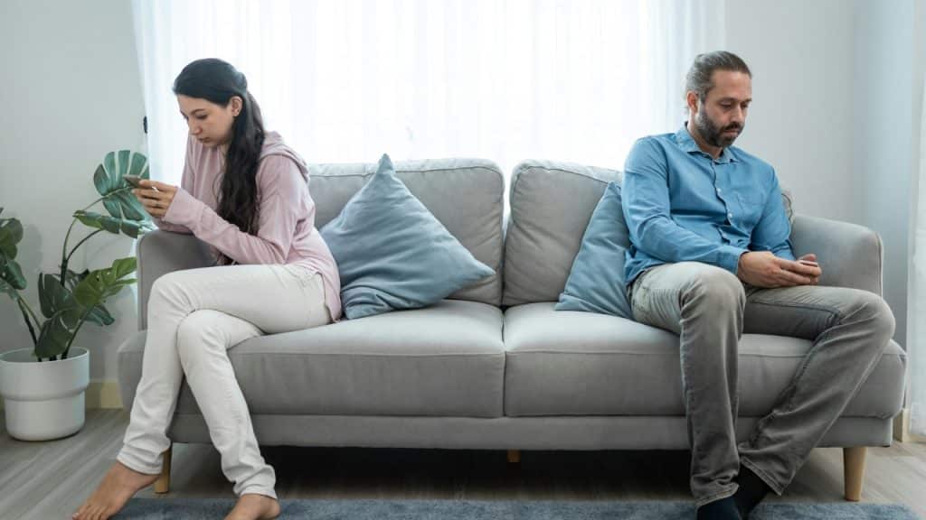 A woman and a man sit on opposite ends of a sofa using their smartphones.