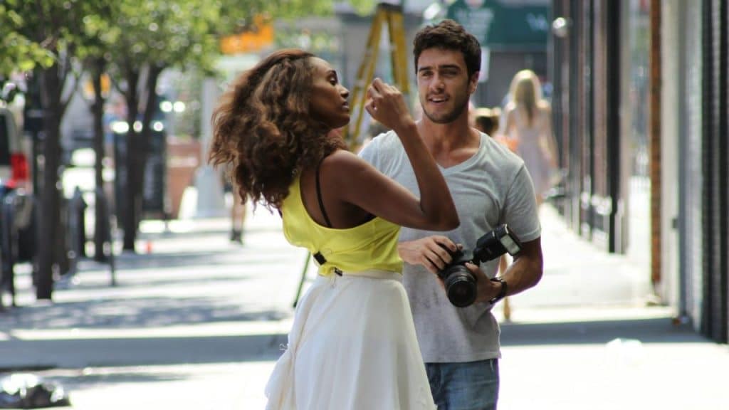 A woman in a yellow top adjusts her hair while a man holds a camera.