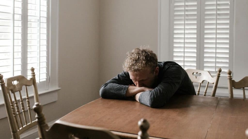 A person with curly hair leaning their head on their arms at a wooden dining table.