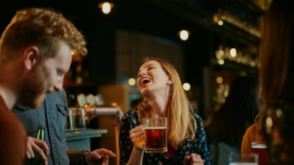 A woman laughs loudly while holding a beer mug at a bar with friends nearby.