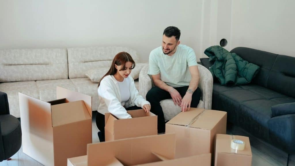 A man and woman sit among several cardboard boxes in a sparsely furnished room.